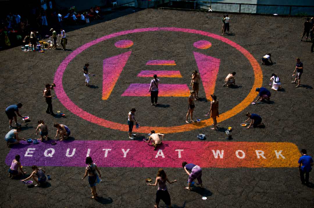 Decine di persone dipingono insieme sul selciato il logo del Pay Equity Pact e la scritta Equity at Work